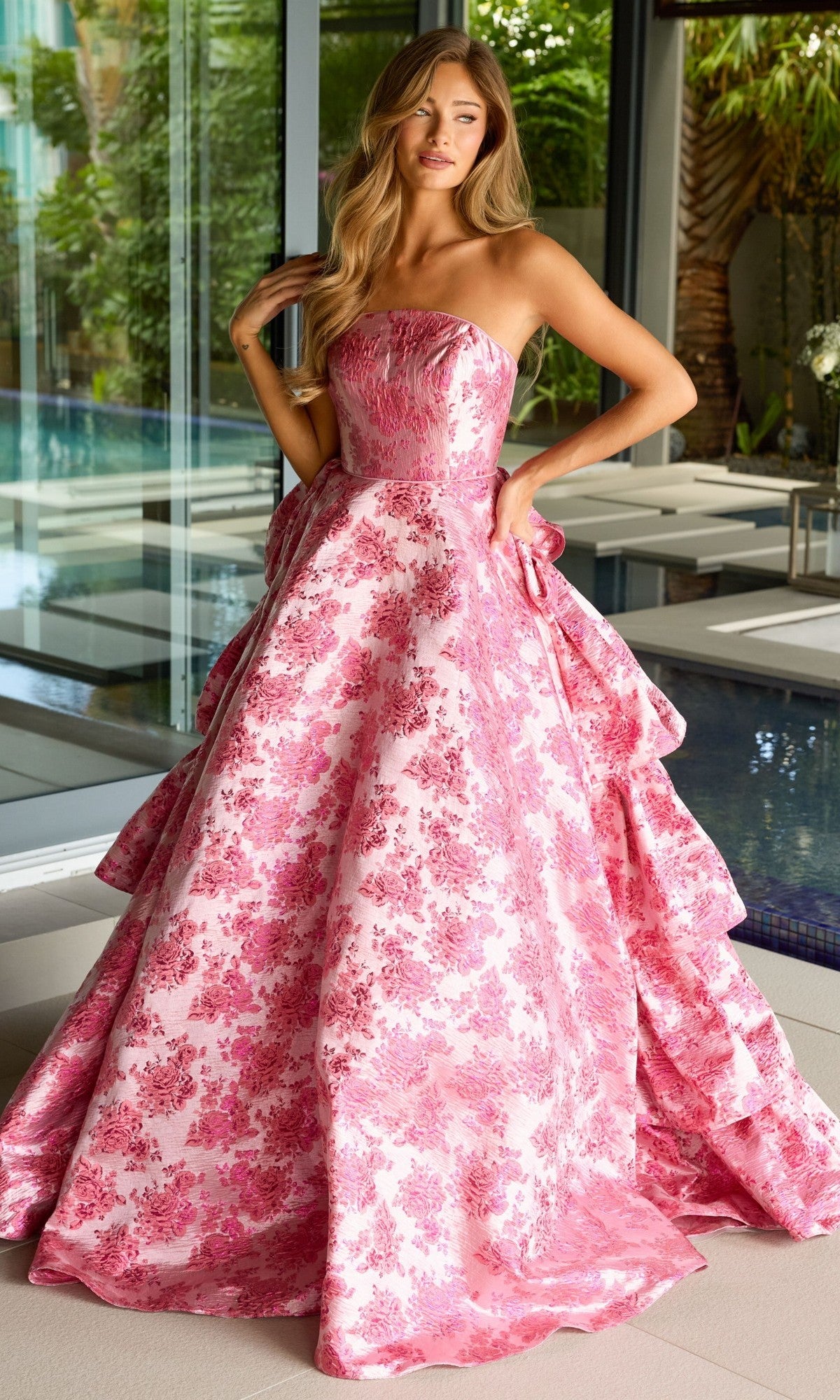 A woman stands indoors in the Ava Presley 26412 Ruffled Strapless Prom Gown, an A-line dress with a voluminous skirt and elegant floral print.